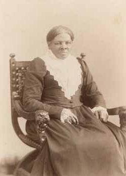 <p>Black and white photograph of a Black woman with her hair parted in the middle and pinned behind her head wearing a black, long-sleeved dress and a triangular lace collar. She sits in a wooden chair with both hands in her lap.</p>