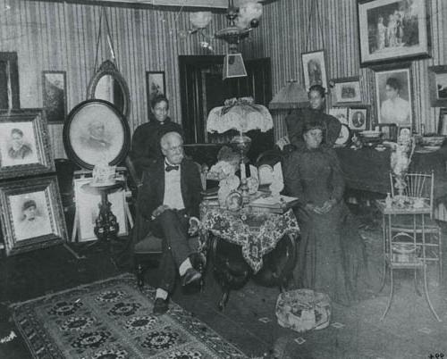 <p>Black and white photograph with a white border depicting a room with striped wallpaper and decorated with several portraits with four people in the center, surrounding a table and lamp. An older Black man in a suit with his legs crossed sits across the table from a Black woman in a black dress. Two other Black women in black dresses stand behind the two people seated on either side of the table.</p>